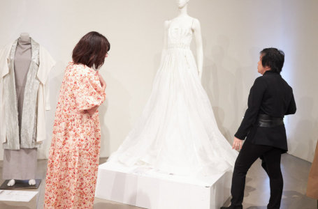 Two women look at a female mannequin wearing a long white dress