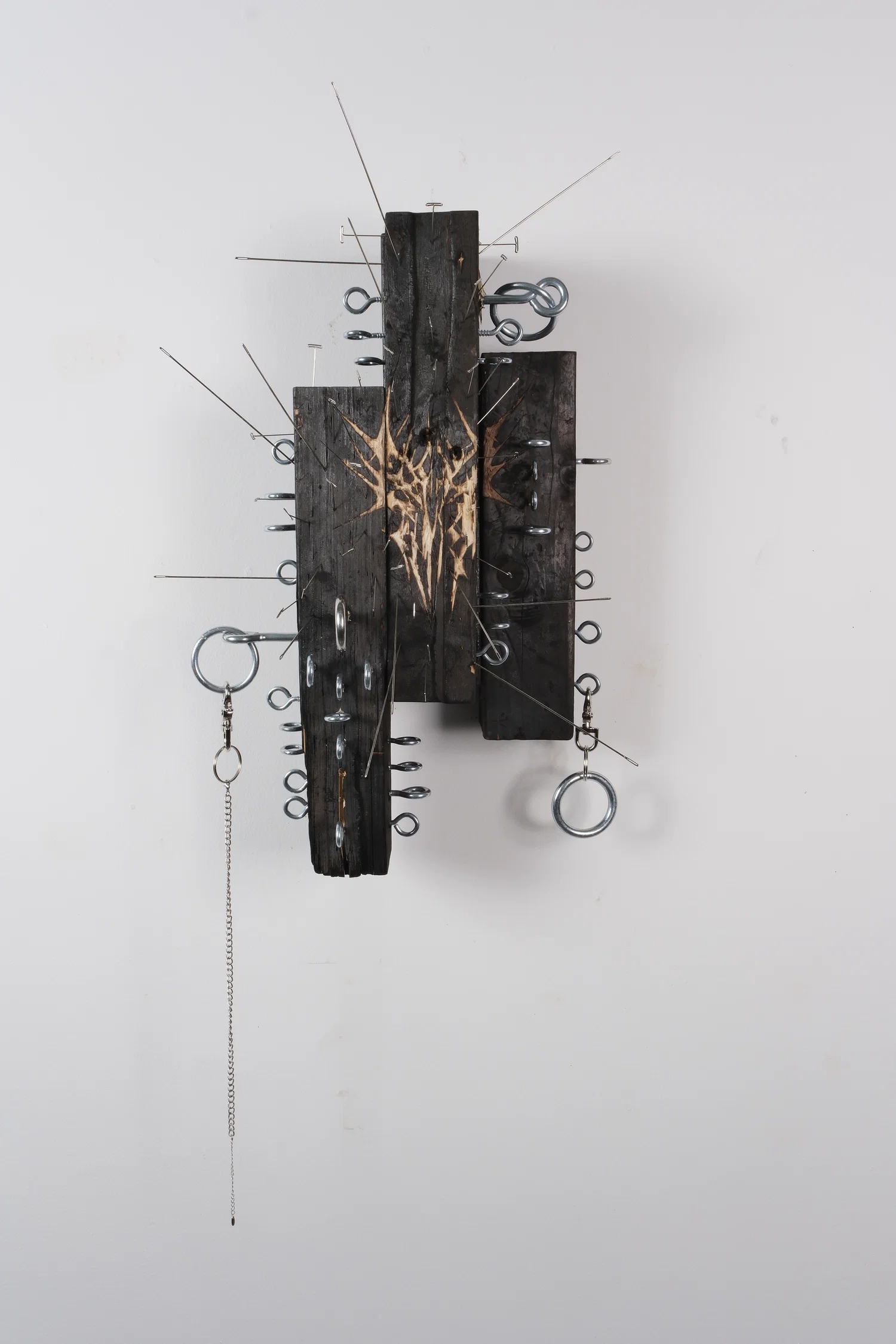 A sculpture comprised of four charred pieces of wood, with needles impaled along the surface, and a sigil carved into the face of the object.