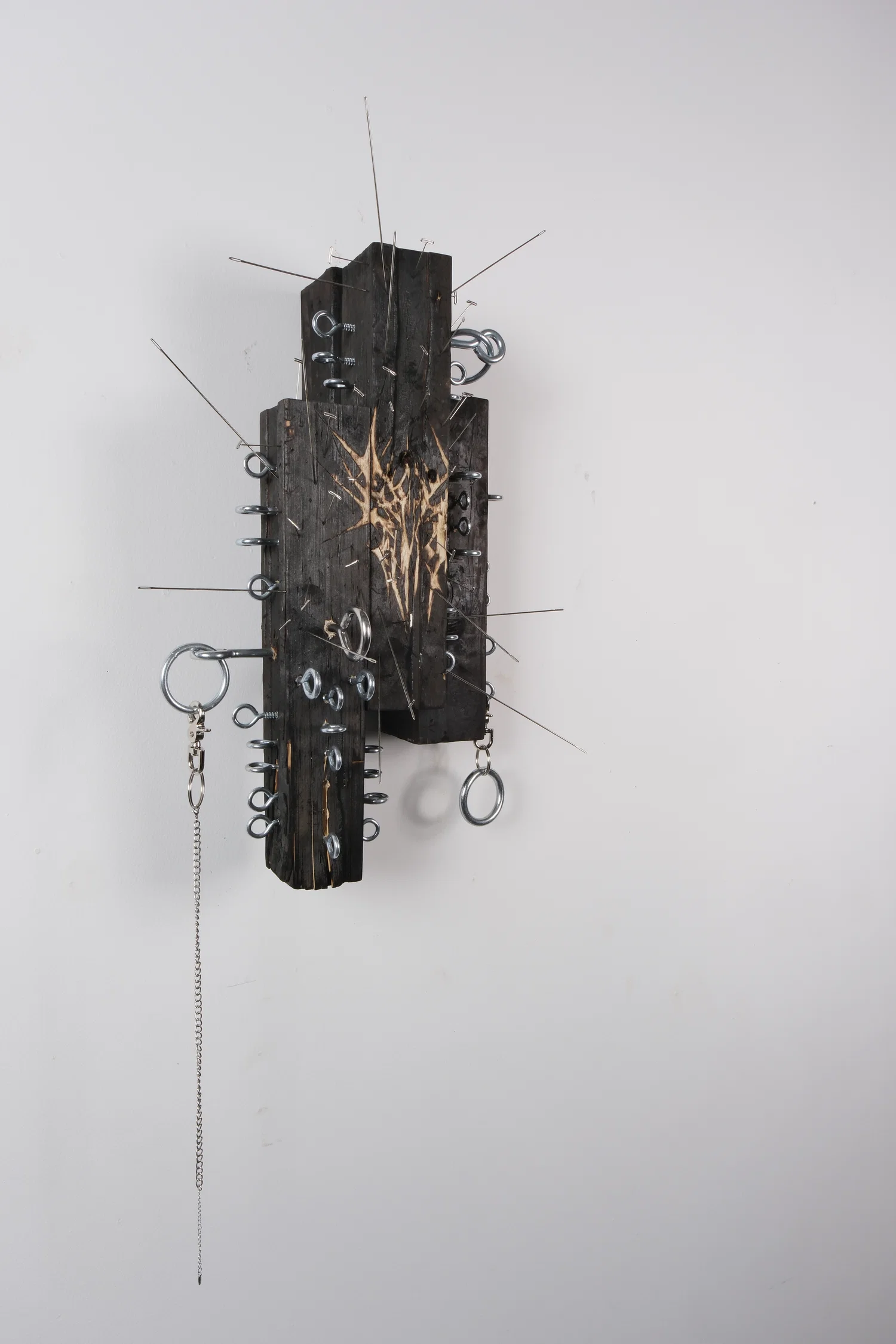 A sculpture comprised of four charred pieces of wood, with needles impaled along the surface, and a sigil carved into the face of the object.