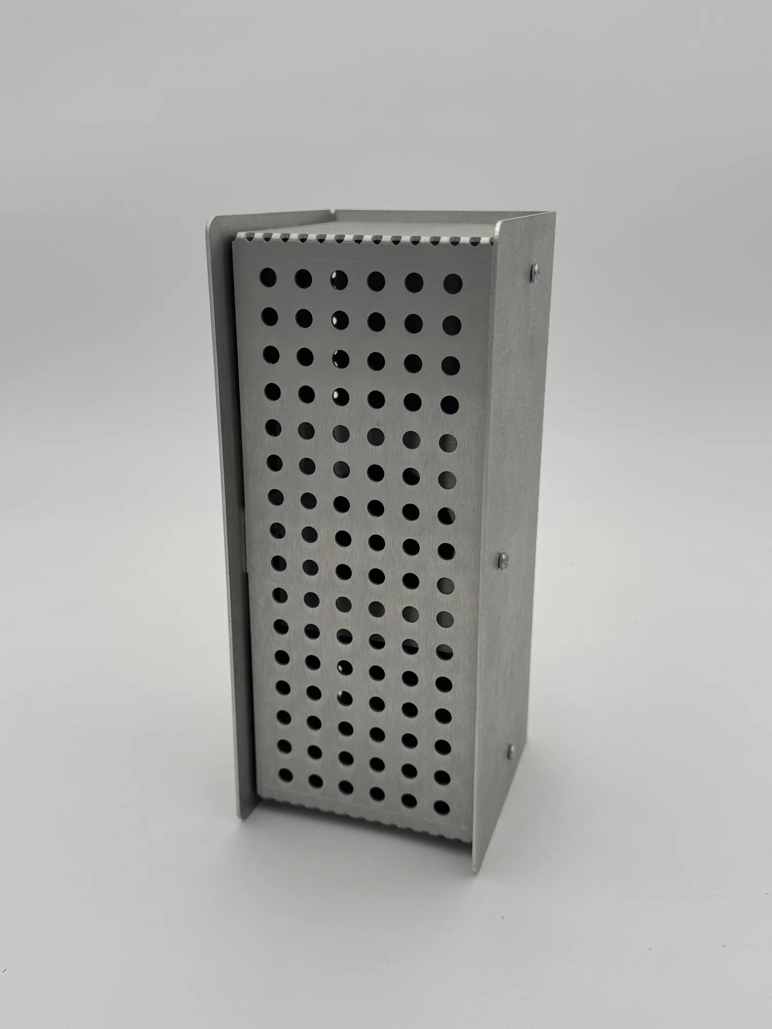 The image shows a compact, rectangular speaker with a minimalist, industrial appearance. The speaker is made of brushed aluminum panels, giving it a sleek, metallic finish. The front face of the speaker features a perforated panel with a grid of evenly spaced round holes, allowing sound to pass through while maintaining a clean, modern aesthetic. The speaker’s body is assembled using small, visible screws on the side panels, emphasizing its modular, easy-to-disassemble design. The overall shape is a simple, upright box with flat surfaces and sharp, clean edges. It has a sturdy, functional look, reflecting a design philosophy focused on durability, simplicity, and repairability. The background is plain and light grey, keeping the focus entirely on the speaker.