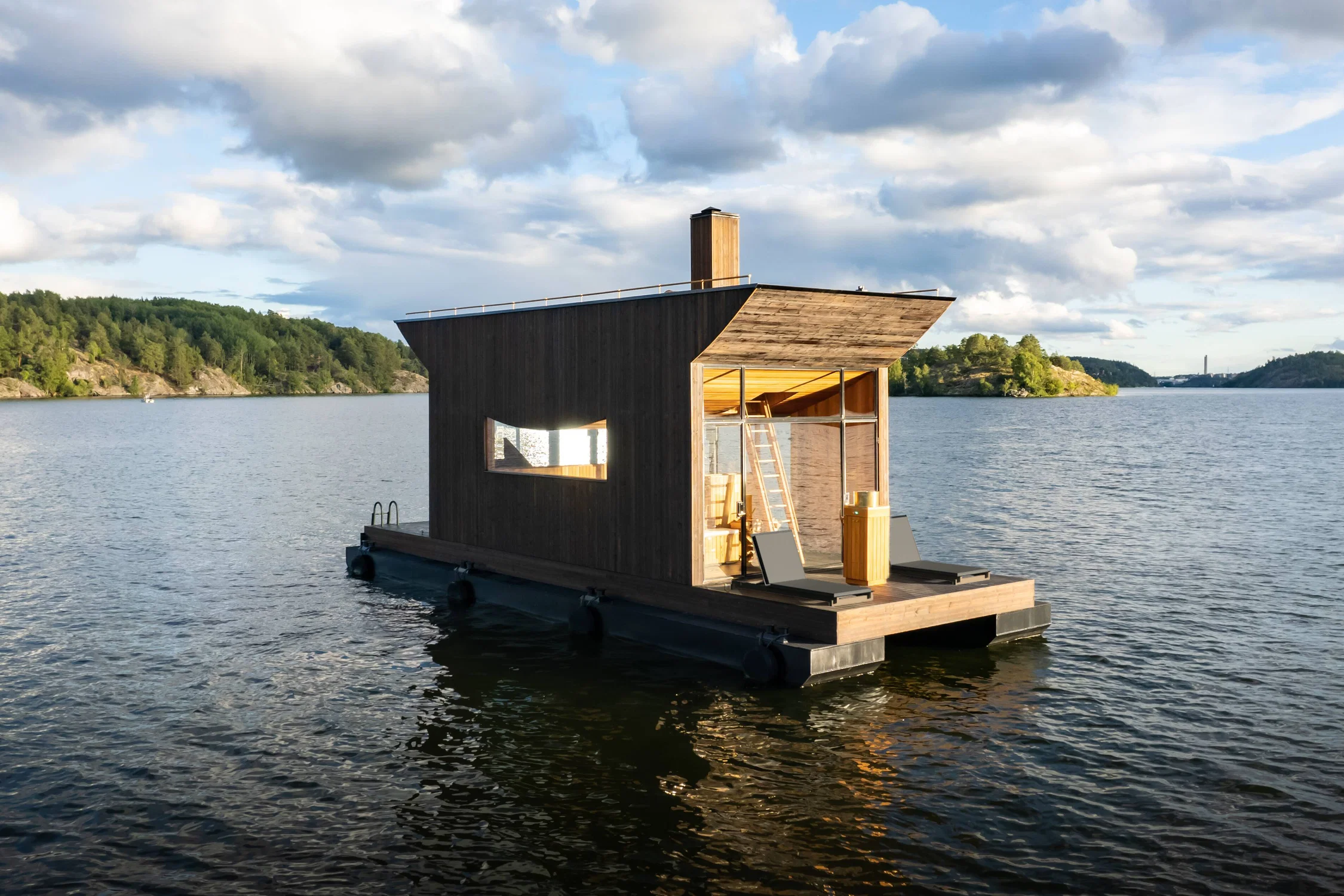 The image shows a small, modern wooden sauna structure floating on a calm body of water, surrounded by forested hills and scattered islands in the distance. The sauna is built on a rectangular floating platform. Its exterior is clad in dark wooden planks, with a slightly angled roof that extends outward, providing some shade to the open deck area in front. On the deck, there are two minimalist lounge chairs — the Vila Lounge Chairs — positioned side by side facing the water. The sauna features large glass doors and windows, allowing views through to the interior where wooden seating and a ladder are visible. The lighting suggests late afternoon or early evening, with soft golden light and scattered clouds in the sky, reflecting gently on the water’s surface.