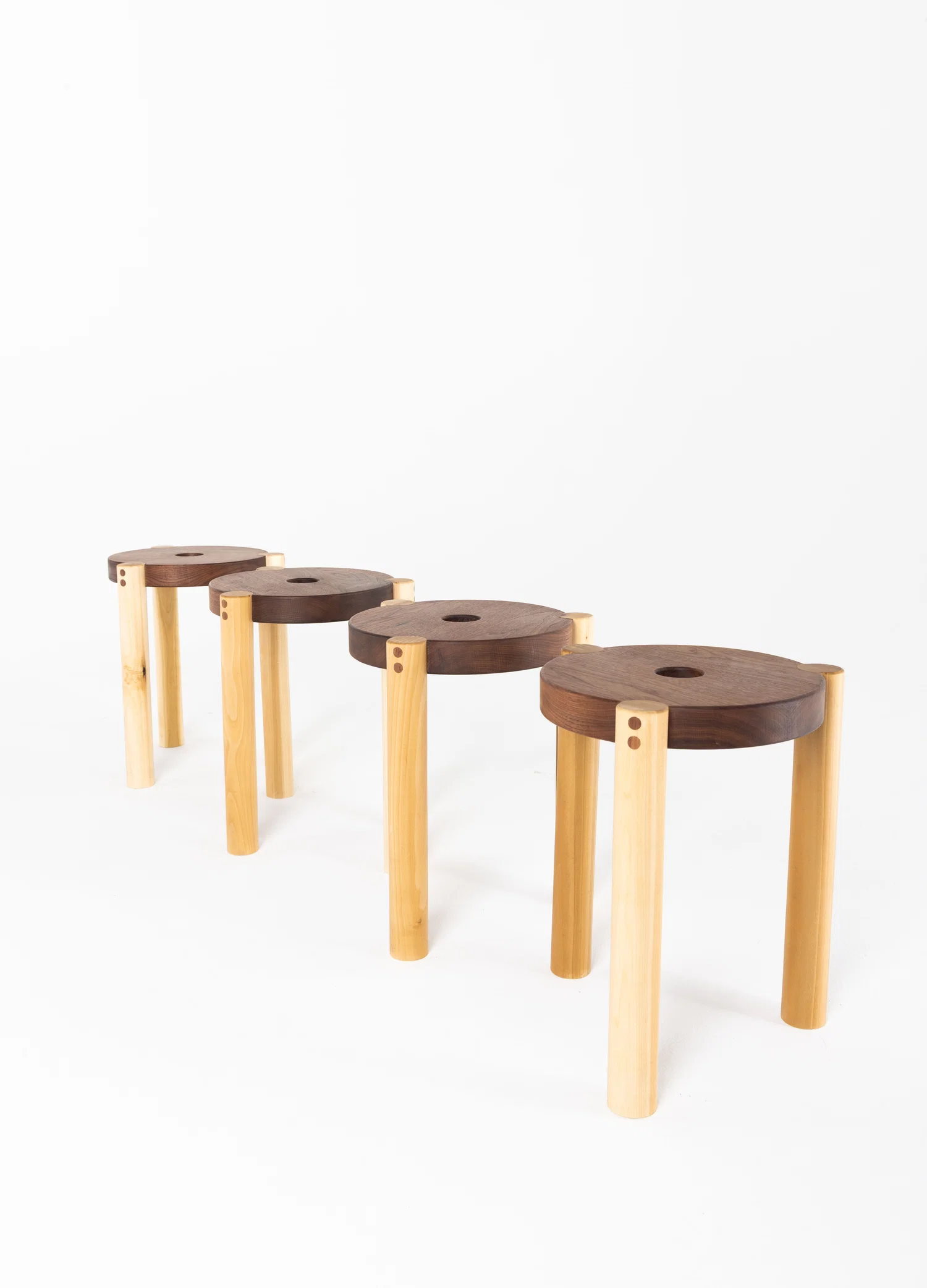 A lineup of four stools of the same design. They have a circular walnut seat with a hole cut out of the center. Three poplar legs featuring two small walnut circle inlays on each leg.