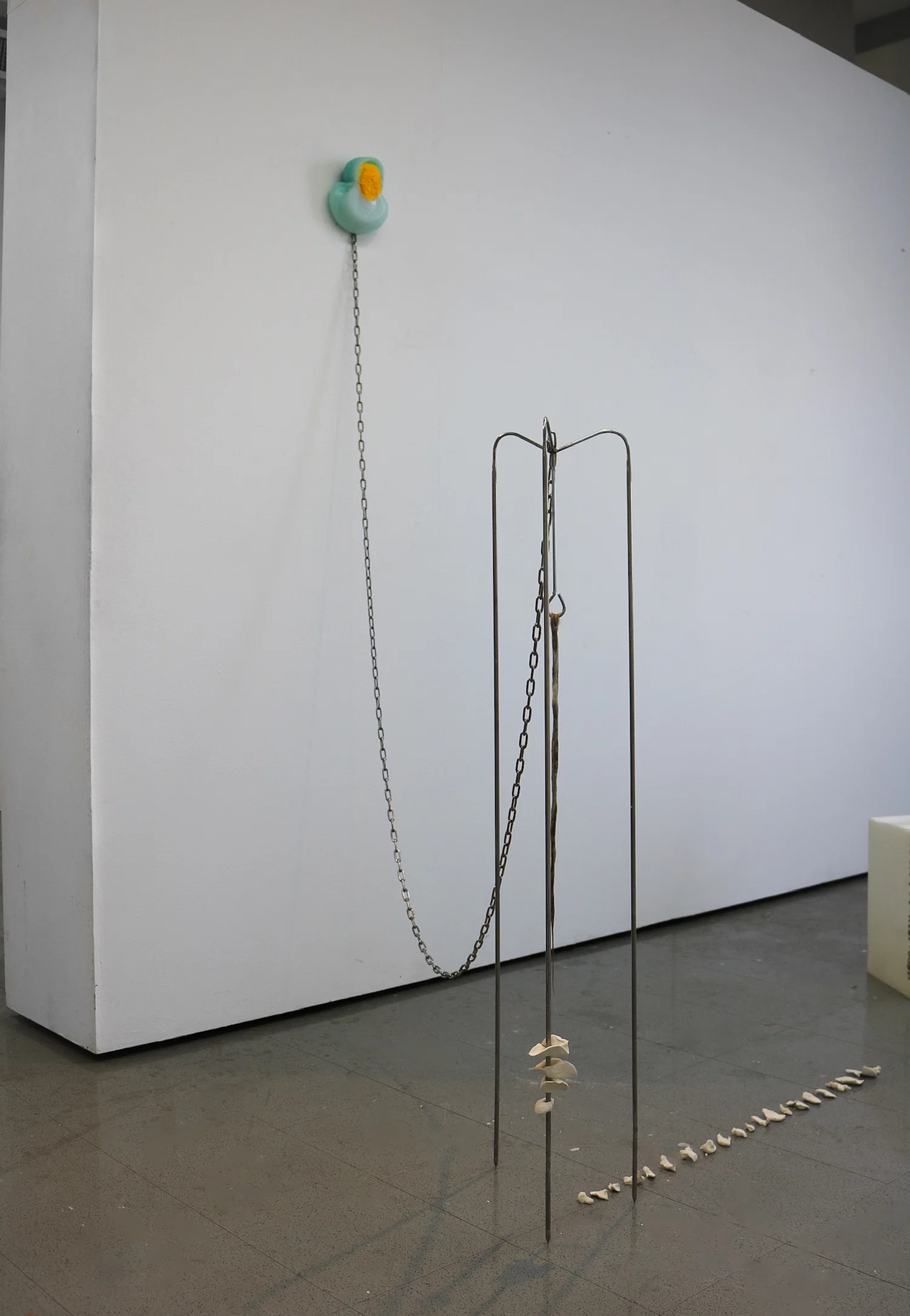 Three pronged talk steel rod sculpture made from a meat hook in its center is a long rope of hair coated with wax, a chain welded to the top of the structure leads to the wall where a small blue and yellow plastic form is chained