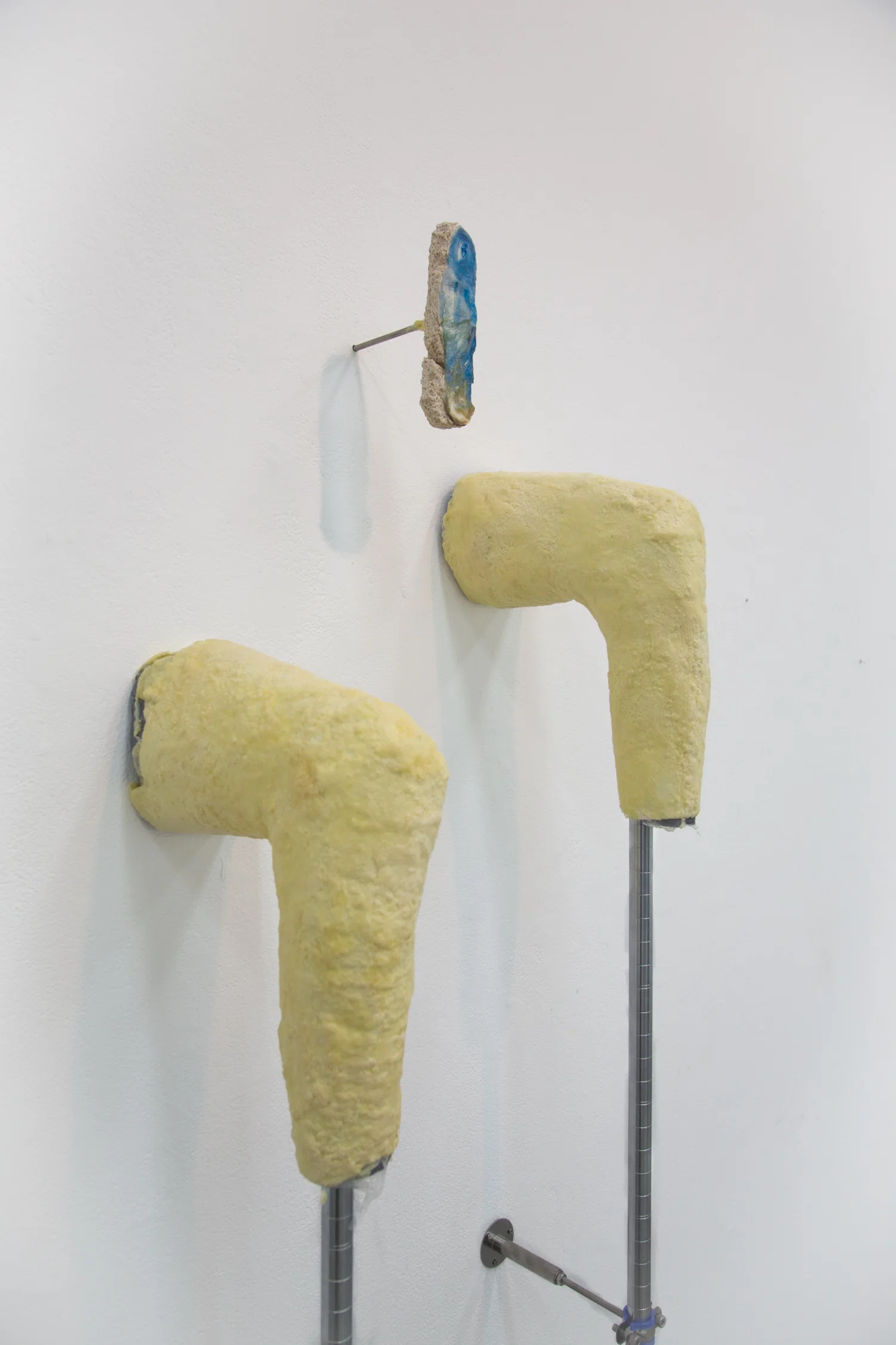 Two wax coated yellow legs jut out of the wall they have stilted metal poles that are clamped onto the wall with pipe fixtures. Above the legs is a fragment of a wall that comes out, with an image of the Virgin Mary digitally edited on top of a doll wrapped in cellophane.