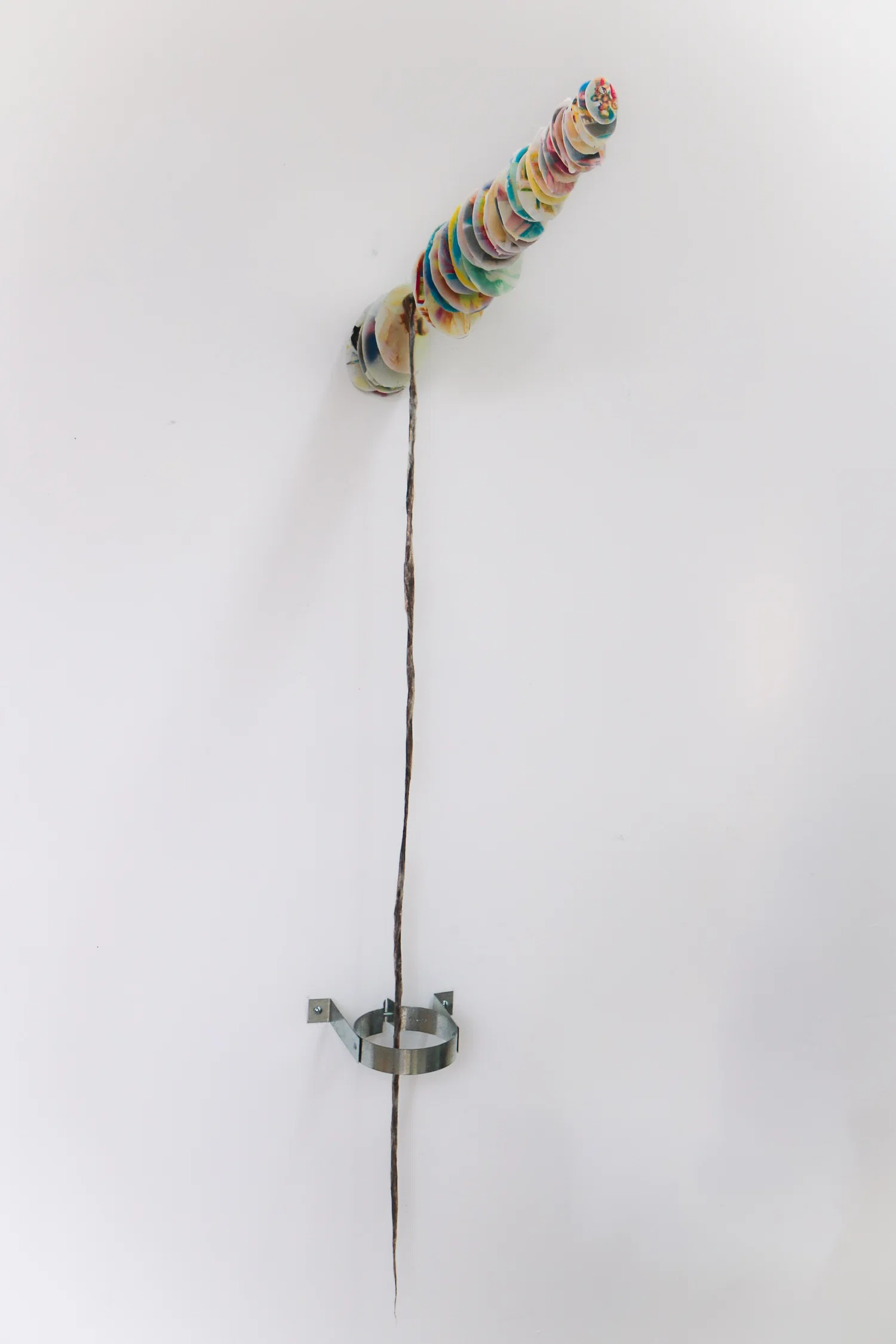A steel rod comes out of the wall. A series of over saturated wax dipped egg shaped photos are lined on the rod. Hanging from the rod is a long wax coated rope of hair. The hair enters an open pipe clamp.