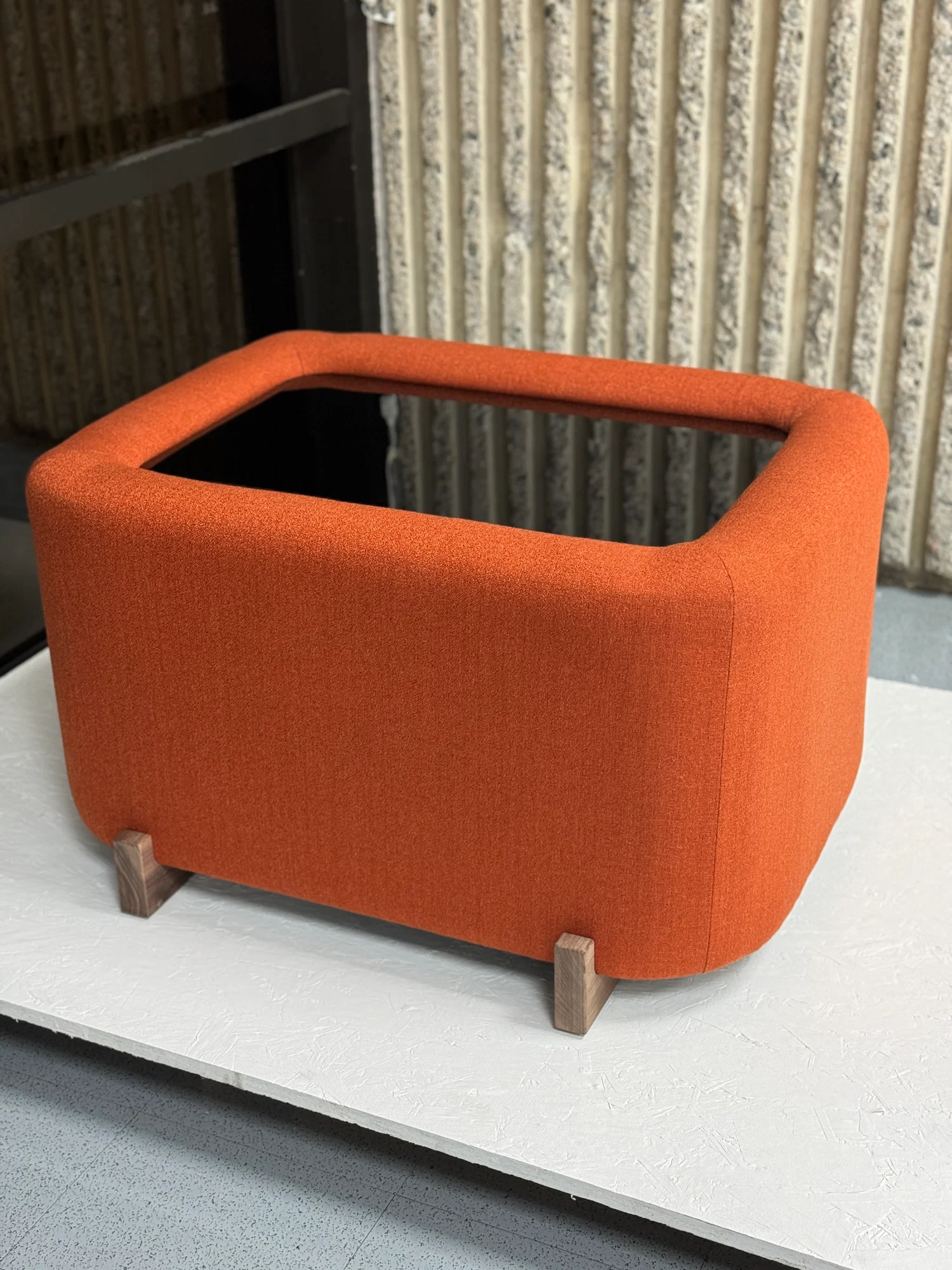A square, burnt orange upholstered furniture piece with rounded edges and wooden legs.