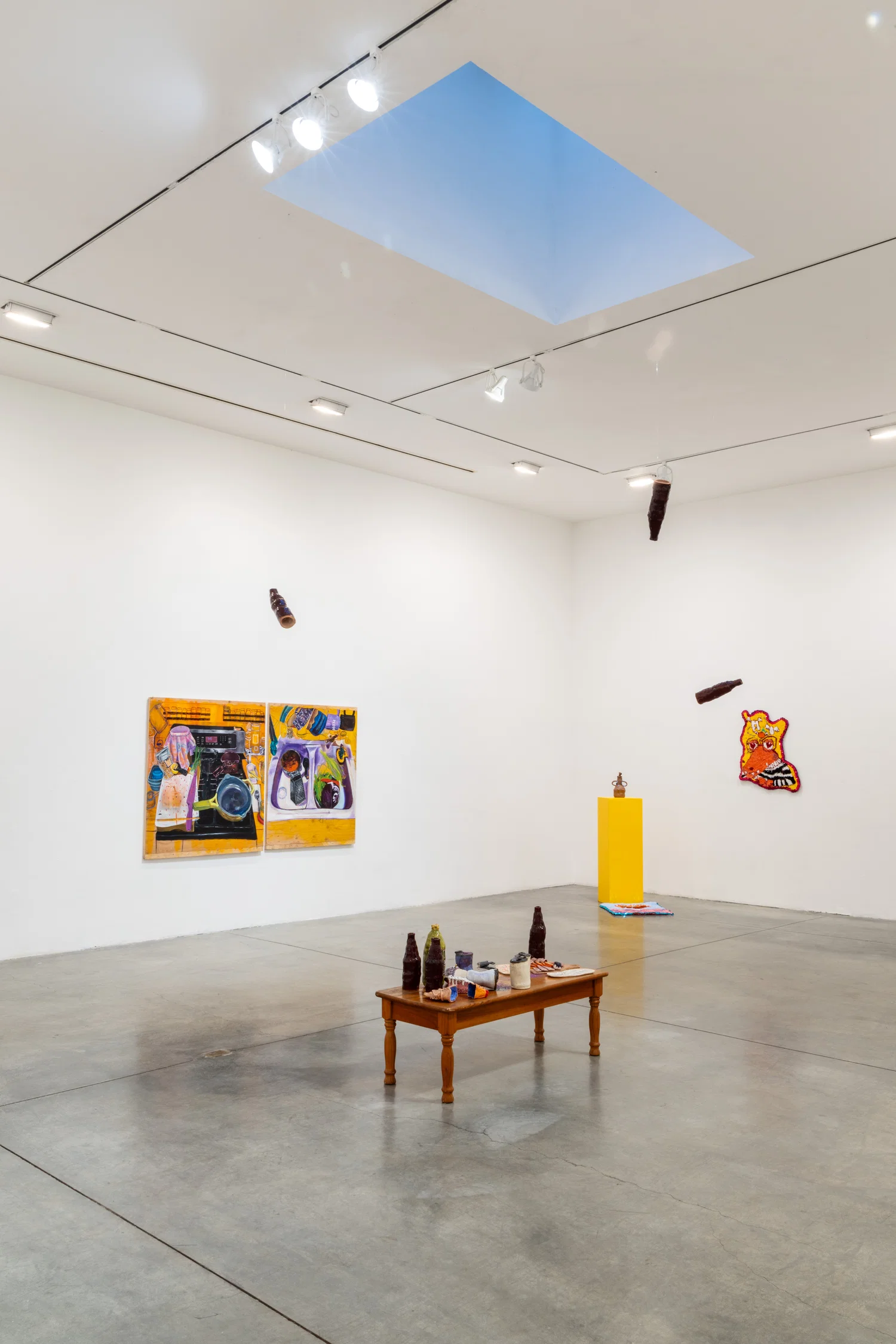A photograph showing a white room, with a short wooden coffee table with small ceramic bottles, cans, and other small food objects in the middle of the room. On the left wall, there is a painting diptych depicting a kitchen stovetop and a kitchen sink. On the right wall, there is a colorful rug with a sequin border. In front of this rug, there is a yellow pedestal with a brown ceramic vase on top and there is a blue rug on the floor in front of the pedestal. There are three ceramic bottles hanging from the ceiling and on the top of the ceiling is a skylight casting a blue shadow.