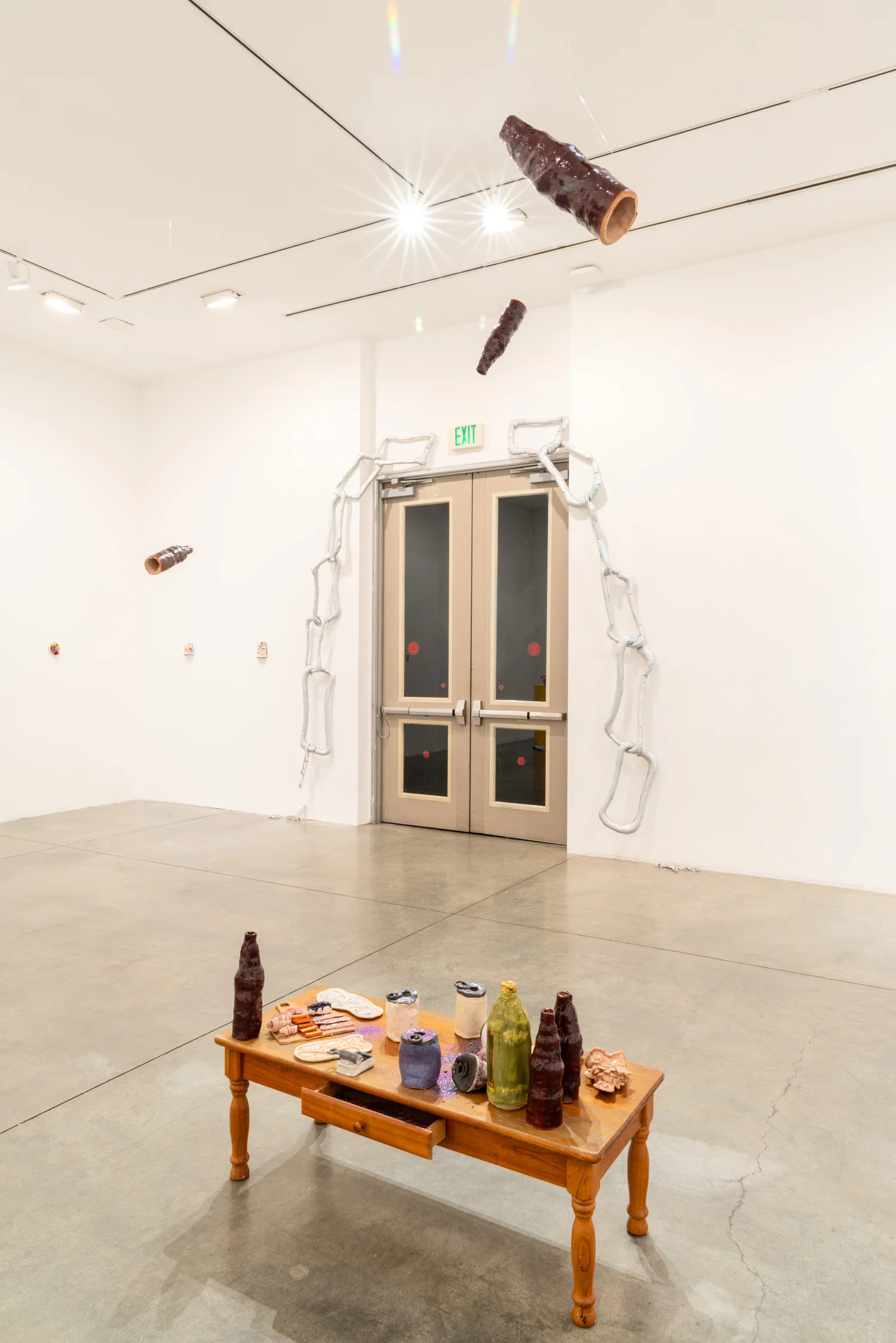 A white room, with a short wooden table with an assortment of small ceramic sculptures and scattered glitter. Hanging from the ceiling are three ceramic bottles. In front of the entrance are silver holographic plushy chains. On the far walls on the lefthand side are small ceramic wall reliefs.