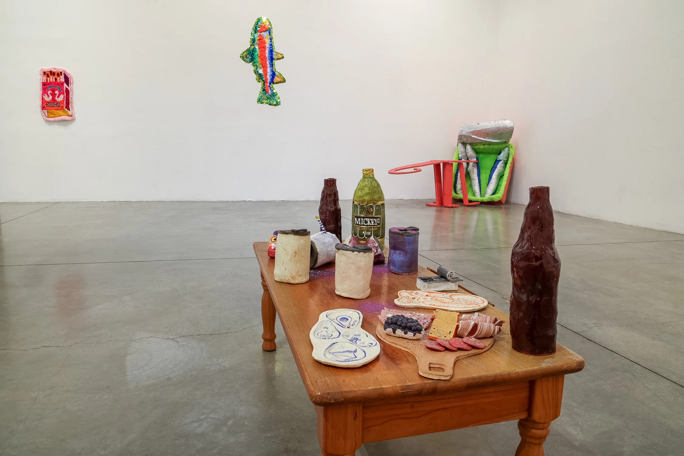 A white room with a pink rug on the far left wall, a tufted fish with sequin border, a pink wooden alligator table sculpture pointed towards a cardboard and fabric sculpture of a sardine can, and a close up of a short wooden coffee table with small ceramic sculptures and glitter scattered on top.