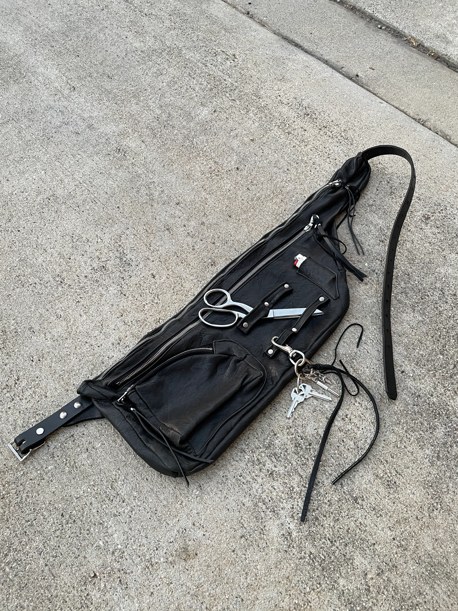The Apron Side Bag is a bag that was designed for a person working in a fashion fitting that needed quick access to their tools. The bag is to be worn around the waist to allow for easy reach of your tool. It’s made of black lambskin leather with 3 metal silver zippers running along the entire bag.