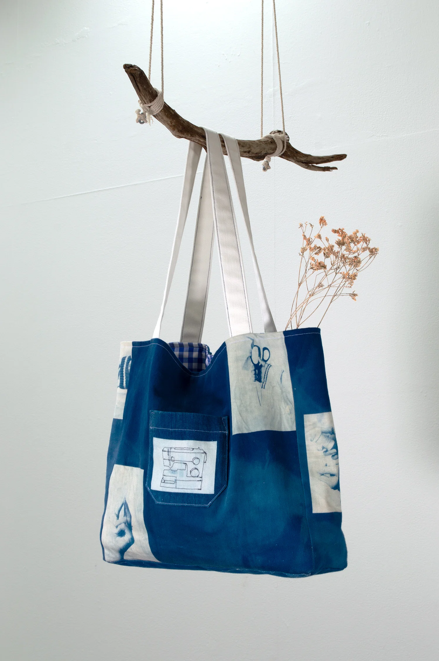 A close up photo of the front of the tote bag hung on suspended branch. Front patch pocket has a sewn image of a sewing machine.