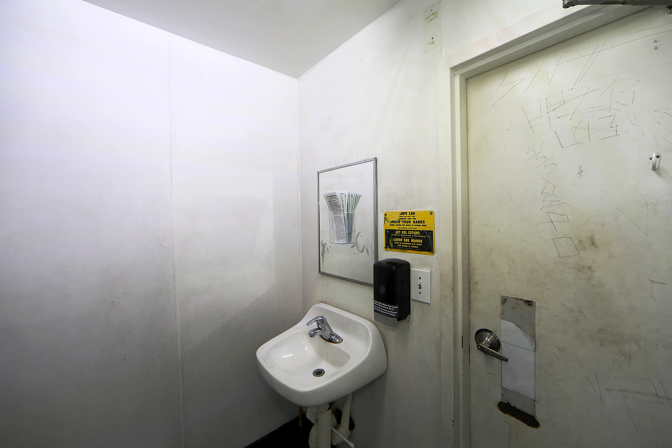 a photograph using a wide angle lens inside a gas station bathroom where an acrylic pamphlet holder has been attatched  the the mirror above the sink, inside the holder is a stack of complaint pamphlets.