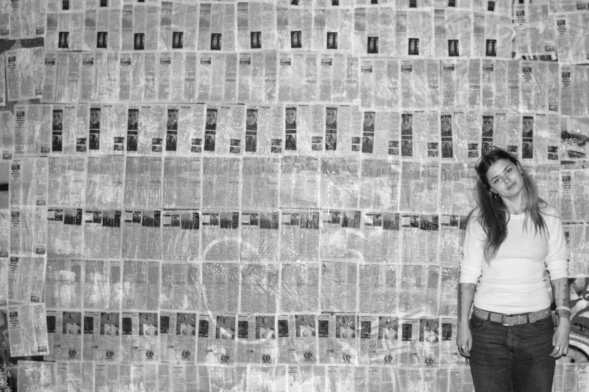 a black and white photograph of me in front of a wall where I have wheatpasted the pamphlets flat to cover the entire wall. they are organized by editions 1-7 starting at the top of the wall and working down.