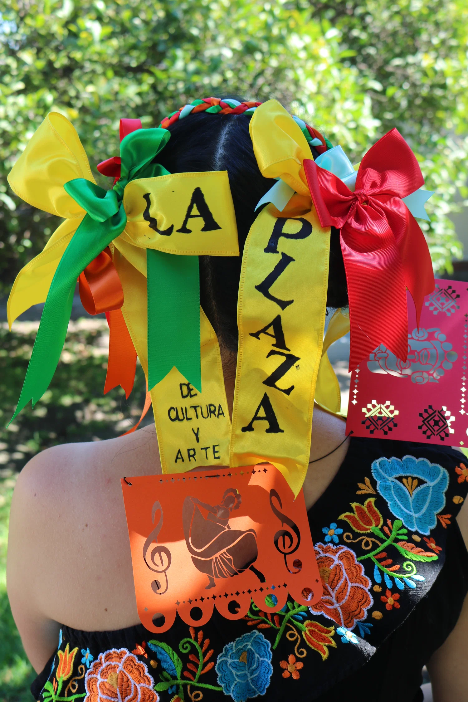 Young woman turned back, with hair braided into a crown braid and ribbons all around her head. Some ribbons are used to spell out LA Plaza de Cultura Y Artes.
