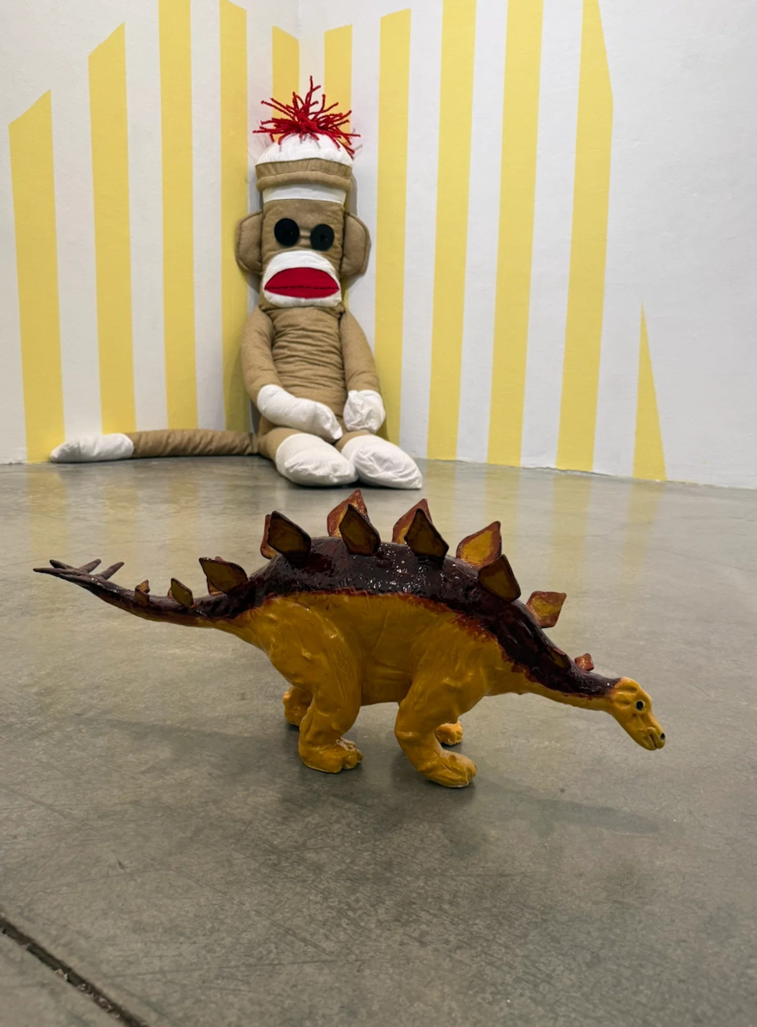 Yellow ceramic Stegosaurus dinosaur and soft sculpture sock monkey leaning up against yellow and white striped wall