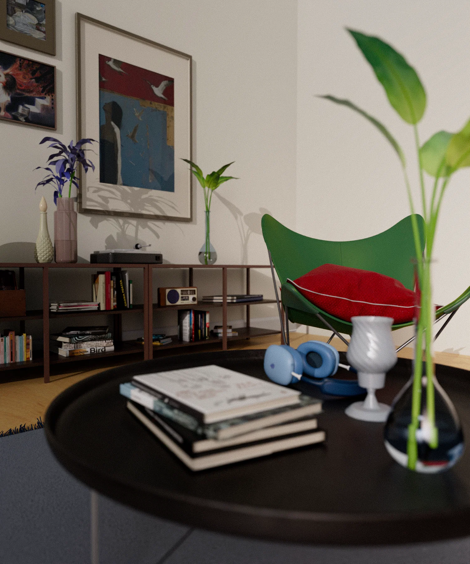 A cozy, eclectic living room corner featuring a dark circular coffee table in the foreground with a stack of books, blue headphones, and a small glass vase. In the background, a green butterfly chair with a red pillow sits next to a low wooden bookshelf filled with books and a vintage radio. The wall is decorated with several framed art pieces, and various potted plants and vases are scattered throughout the space, creating a lived-in, artistic atmosphere.