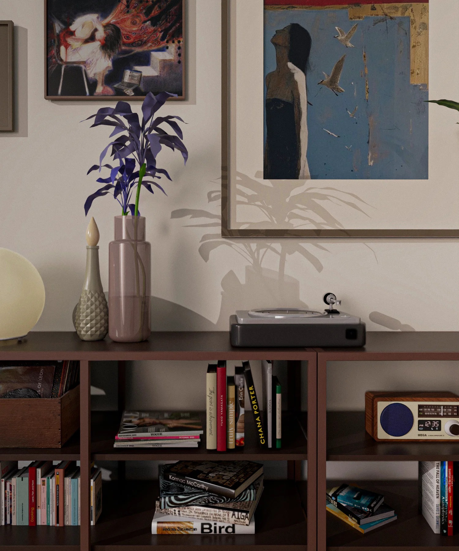 Alt Text A close-up shot of a dark wood shelving unit topped with decorative items. On the top surface sits a record player, a pink glass vase with purple leaves, a textured ceramic bottle, and a round lamp. The shelves below are filled with various books, magazines, and a vintage-style wooden radio. Framed art pieces hang on the wall above, with the shadow of the purple plant cast softly against a large print of a woman and birds.