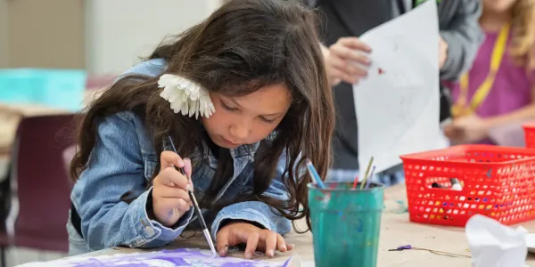 A female Summer Youth Camp student with a white flower in her hair works on her painting