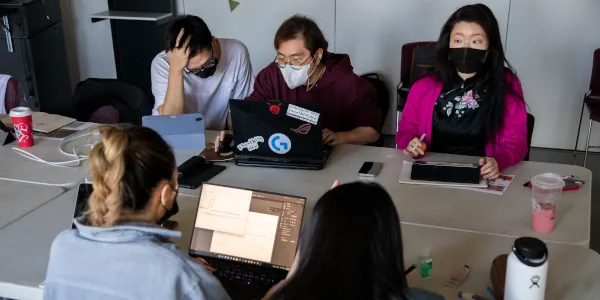 Five students sitting at a large table and working on their laptops and tablets
