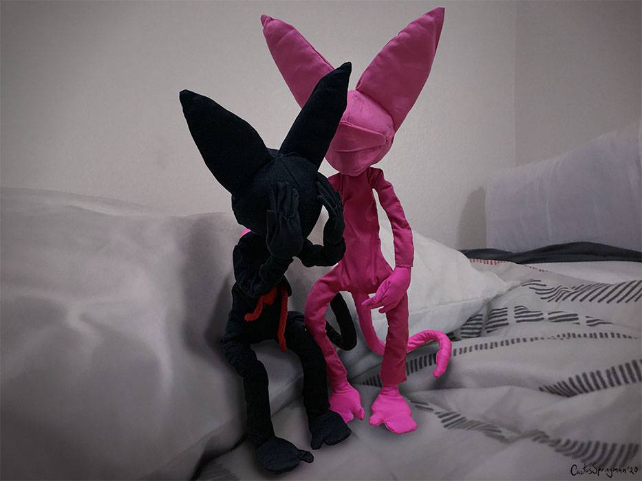 Please Don’t Cry Brain Cats, by Cactus Springman. A photographic print of two soft sculptures, a black cat and pink cat. The black cat is sitting on a pillow, head in their hands, while the pink cat tries to comfort them. Both cats are on top of a human-sized bed.  